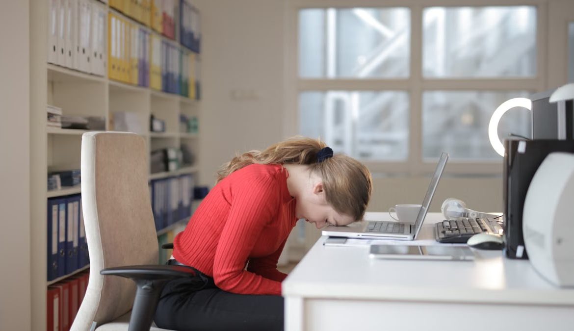 Tired woman in red sweater naps on office desk beside laptop, overwhelmed by remote work.