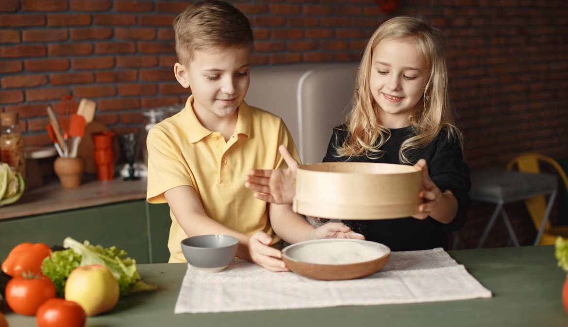 Content little friends in casual wear sifting flour through wooden sieve into round plate put on towel on table with fresh vegetables in loft style kitchen at home